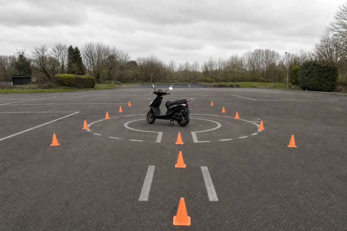 Piste de plateau d’auto-école avec plots orange où un scooter 50 cm³ s’exerce au freinage, sol bitumé